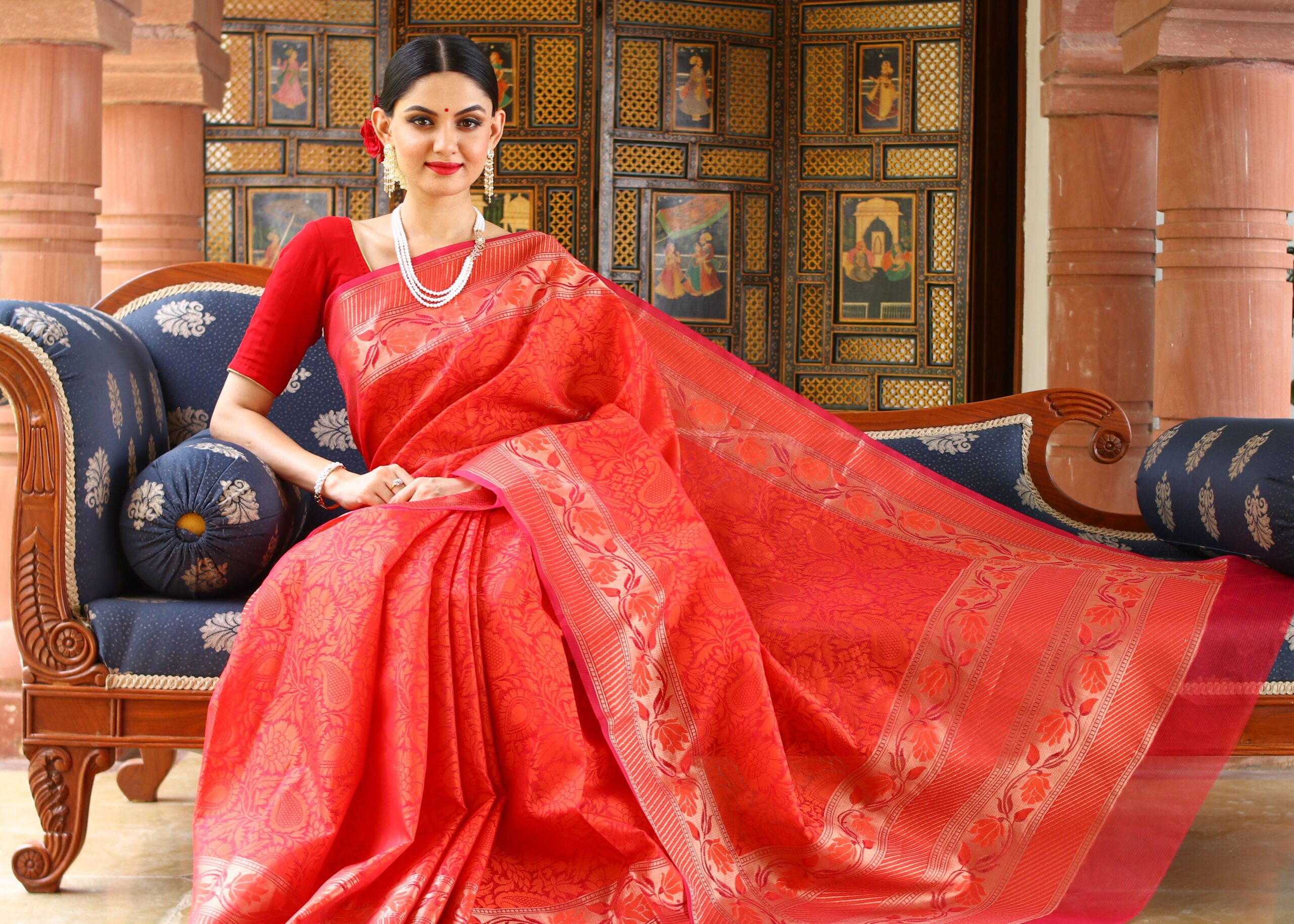 Woman in red saree poses elegantly indoors, highlighting traditional fashion and culture.